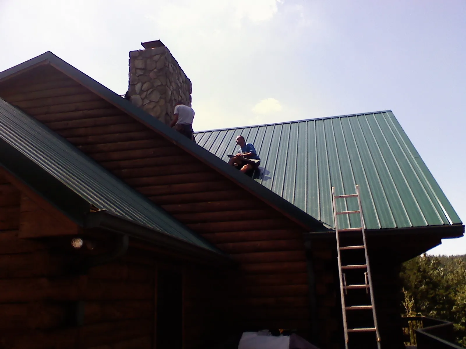 Roofers installing green metal panels beside a stone chimney on a log home in Spring City